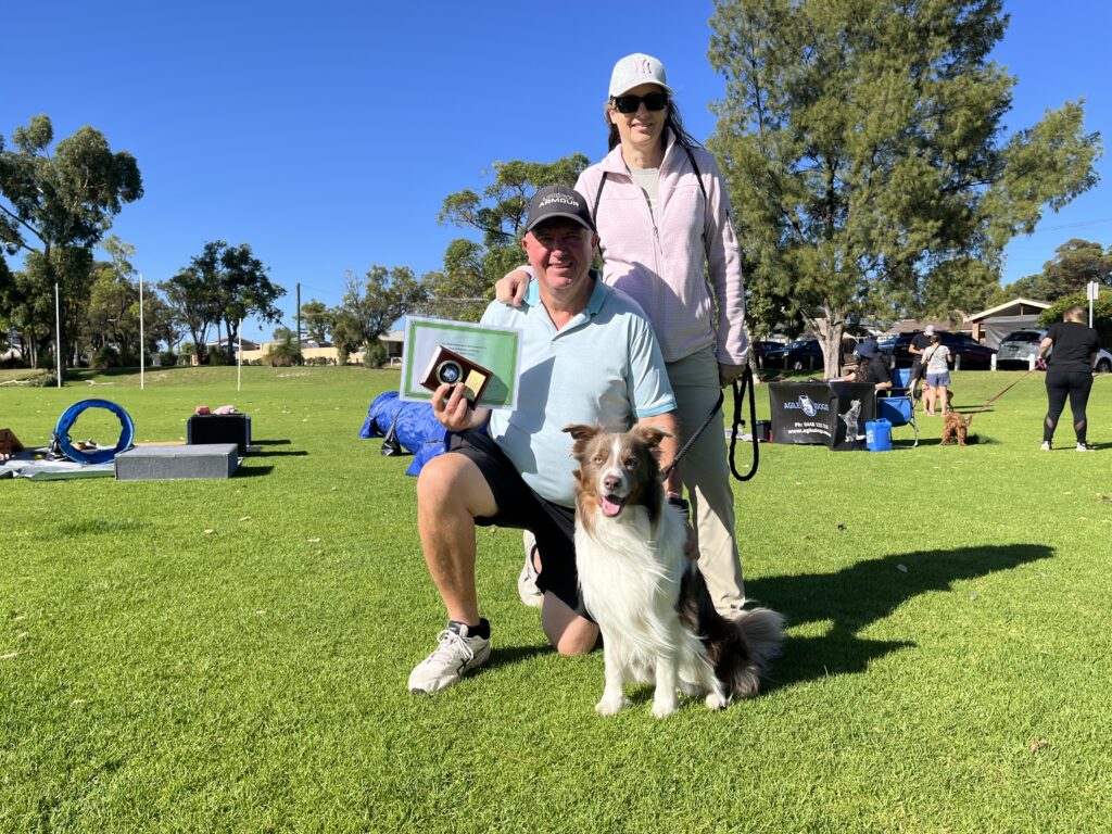 K9 agility competition winners with their dog and award on a sunny day in a park.