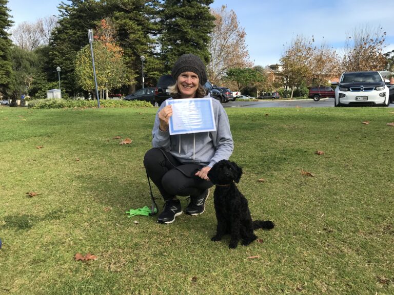 Woman with a black poodle holding a training certificate outdoors.