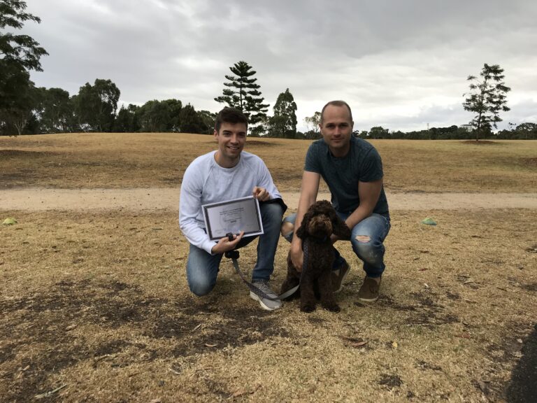 Two men with a dog outdoors, celebrating training achievement at Agile Dogs.