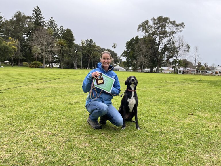 Woman with her dog holding a medal in a park setting.