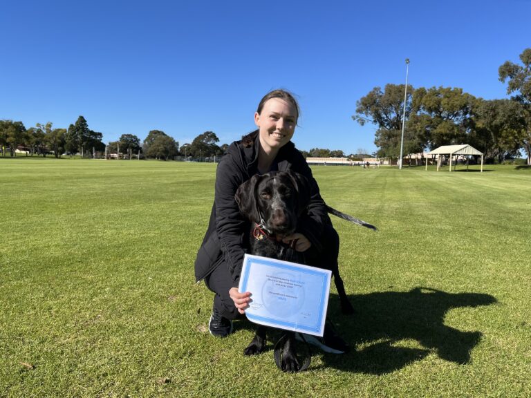 Woman with a black guide dog holding a certificate outdoors in a park.