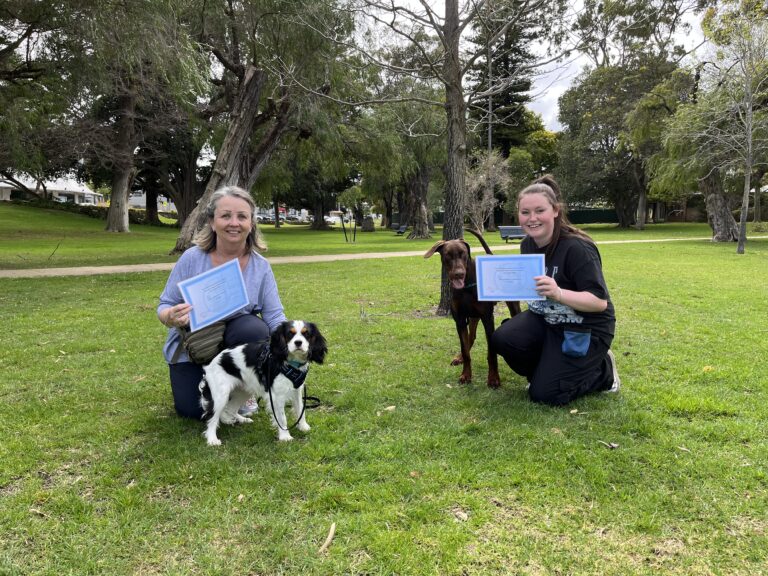 Two women with dogs holding certificates in a park setting.