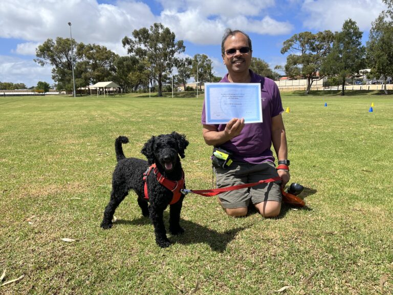 Man kneeling with black poodle holding a certificate outdoors.