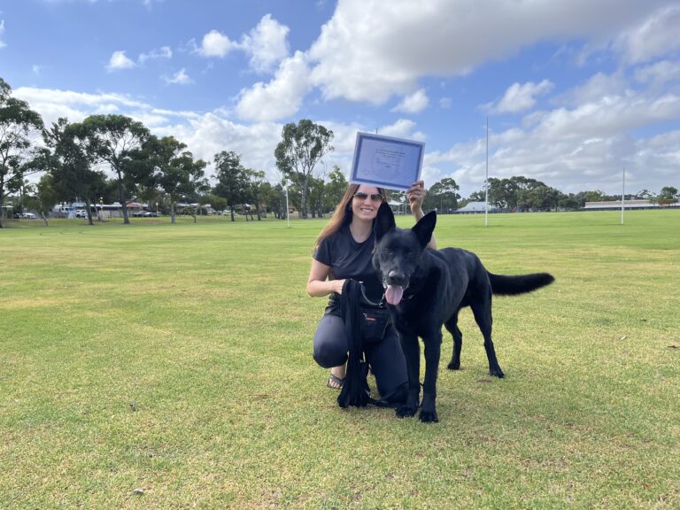 Woman with black dog holding training certificate outdoors on a grassy field.