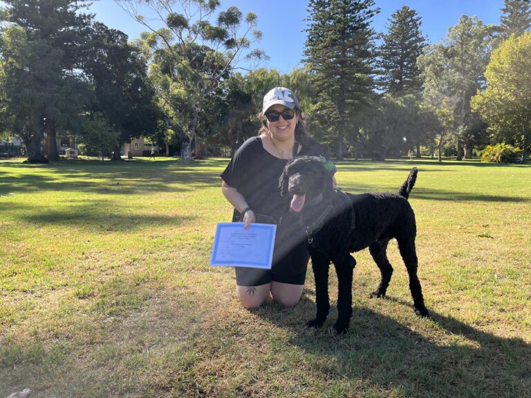 Dog training certification celebration at the park with a smiling woman and her black dog.