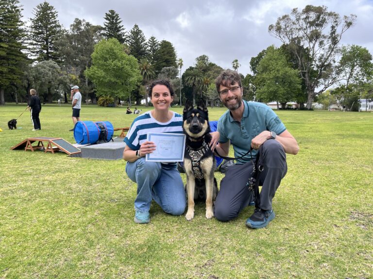 Happy dog trainers celebrating with a certified dog in a park setting.